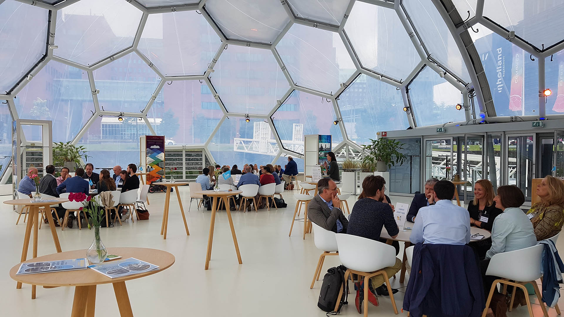 Duurzaamheidsstratiegieën voor je bedrijf. Ken je deze al? Een blog van PR Goeroes, hét pr-bureau dat media-aandacht en zichtbaarheid voor jou en jouw bedrijf genereert. Op de afbeelding zie je mensen in overleg aan ronde tafels onder een ronde glazen koepel.