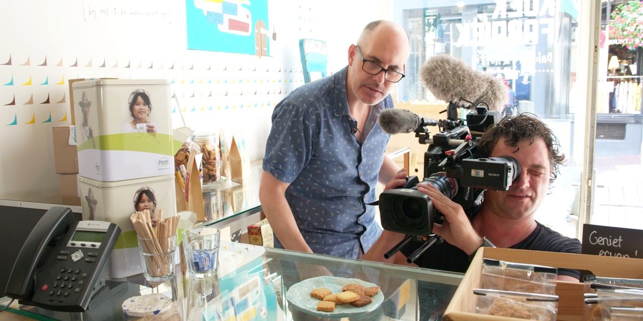 Jouw bedrijfsverhaal op een (gratis) LinkedIn-video? Video is het medium waar mensen op social media de meeste tijd aan besteden. Hiermee is het een hele krachtige marketingtool voor zakelijke dienstverleners. Onze partner Verhaalmakers wil graag meer ondernemers helpen met het inzetten van video. Afgebeeld staat een witte cameraman die een schotel met koekjes op een toonbank filmt, schuin achter hem kijkt een man in een blauw overhemd met korte mouwen met hem mee.